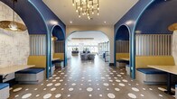 Located at 1010 E Orange St in Tempe, Arizona, this generously sized single-floor amenity space delivers a dynamic, design-forward environment that seamlessly blends social and recreational functions. The entry corridor makes an immediate impression with bold cobalt blue arched alcoves housing cozy booth seating, a polka-dotted concrete floor, and a sculptural brass chandelier overhead. Moving deeper into the space, the open-plan layout reveals distinct yet interconnected zones. A lounge area features plush swivel chairs, curved blue sofas, a large-screen TV, and colorful vertical slatted wall installations. A centrally positioned billiards table anchors the entertainment zone, surrounded by additional seating nooks accented with tall indoor plants. A bar-style counter with white barstools sits beneath a warm wood ceiling panel and a vivid, backlit shelving display in rainbow hues. The pool-facing wing boasts soaring double-height windows and glass sliding doors that frame views of a resort-style pool and palm trees, flooding the interior with abundant natural light. Herringbone-patterned wood flooring, hanging egg chairs, and a striking abstract line-art feature wall add to the eclectic, artistic character of this thoughtfully curated communal space.