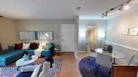 This single-floor residence showcases an open-concept layout that seamlessly connects the living and dining areas, creating a cohesive and welcoming environment. The living area features warm hardwood-style flooring underfoot and is anchored by a vibrant teal sofa accented with colorful throw pillows, complemented by layered area rugs that add texture and visual interest. A large abstract artwork adorns the wall above the sofa, while an adjustable floor lamp provides focused ambient lighting. The space flows naturally toward the front entry, where a classic six-panel white door serves as a focal point. A textured ceiling with a visible HVAC vent adds to the practical detailing throughout. An elegant arched doorway transitions into the dining area, which features a round glass-top table surrounded by upholstered chairs, all set atop a deep purple circular rug. Tall floor lamps in both areas provide warm, diffused lighting. The dining space also benefits from additional overhead track lighting. Soft gray walls throughout maintain a calm, neutral backdrop that allows the colorful furnishings to stand out, while the consistent flooring ties the entire space together into one harmonious living environment.