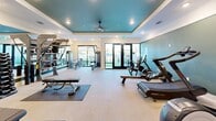 This fitness center is a thoughtfully designed, single-floor workout facility offering a comprehensive exercise environment. The space is distinguished by its striking tray ceiling, painted in a bold teal-green hue and fitted with recessed lighting and a ceiling fan, creating a modern and energizing atmosphere. Crisp white walls with teal-green accent walls complement the contemporary aesthetic throughout.

Natural light flows generously into the space through large windows and a set of glass French doors that open directly to an outdoor area, providing both ventilation and a seamless indoor-outdoor connection. The light-toned flooring adds to the bright, open feel of the room.

The fitness center is comprehensively equipped for a full-body workout. Cardio equipment includes treadmills, elliptical trainers, a StairMaster, stationary bikes, an Assault Fitness air bike, and a rowing machine. Strength training is well-represented with a multi-station cable machine, adjustable weight benches, a dumbbell rack with a full set of free weights, and wall-mounted accessory storage. Protective rubber floor mats are positioned throughout the workout zones.

The layout is open and spacious, allowing for easy movement between equipment stations. The combination of natural light, modern design, and diverse equipment makes this fitness center a functional and inviting space for users of all fitness levels.
