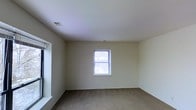 This single-story space presents a straightforward, functional layout centered around a well-proportioned room finished with wall-to-wall carpeting in a neutral tone. The walls are painted in a soft, off-white hue that contributes to a clean and bright atmosphere throughout the interior. A double-hung window positioned along the far wall is fitted with horizontal blinds, allowing for adjustable natural light and a view of the outdoors. The window serves as the room's primary light source, drawing in ample daylight that enhances the openness of the space. A ceiling-mounted air vent indicates a central HVAC system, contributing to year-round climate comfort. The smooth, white ceiling is unadorned, maintaining a simple and uniform aesthetic. On the left side of the room, a partial architectural element — likely a window sill or shallow ledge — adds a subtle structural detail to the otherwise minimalist design. The overall ambiance is understated and versatile, making the space well-suited for a variety of uses. The neutral finishes and uncluttered design provide a blank canvas ready for personalization.