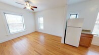 This single-floor unit is designed with a practical and consistent aesthetic, highlighted by warm-toned hardwood floors that run throughout every room. The open layout connects a generously sized living area to the kitchen through an arched passageway, creating a natural flow between spaces. The living area features a ceiling fan and a window that allows natural light to brighten the room.

The kitchen is well-equipped with warm wood cabinetry, granite-style countertops, a gas range, dishwasher, and refrigerator. A window above the counter adds additional natural light to the cooking space.

Multiple bedrooms are present, each outfitted with ceiling fans and windows with blinds. Several bedrooms include closets—some with sliding mirrored doors and others with built-in shelving and hanging rods. A cast-iron radiator appears in select rooms, indicating a classic heating system. The bathroom features a walk-in shower with white subway tile walls and mosaic tile flooring, accompanied by granite-topped surfaces and clean white fixtures.

Overall, the unit presents a clean, functional living environment with classic architectural details and consistent finishes throughout.