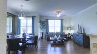 This single-floor residence showcases an open-concept design that seamlessly connects the living and dining areas into one cohesive, well-appointed space. Rich hardwood flooring flows throughout, adding warmth and continuity to the layout.

The living area is anchored by a plush dark brown sofa set adorned with decorative teal and patterned throw pillows, complemented by a sleek glass-top coffee table. A ceiling fan with an integrated light fixture provides both comfort and illumination, while a coordinating accent chair adds extra seating. A side table with a classic table lamp rounds out the sitting area.

Adjacent to the living space, the dining area features a dark round table surrounded by upholstered chairs, set beneath a stylish hanging light fixture. Lush indoor plants add a natural, refreshing element to the room.

Large windows dressed with blue grommet-top curtains allow generous amounts of natural light to fill the space, creating a bright and airy atmosphere. Crown molding trims the ceiling, adding a polished architectural detail, while a textured ceiling finish adds subtle depth overhead. The overall ambiance is inviting and thoughtfully designed, balancing comfort with refined style throughout the open living environment.