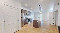 Located on Daro Drive in Herriman, Utah, this two-story residence showcases a clean, modern design with thoughtfully selected finishes throughout. The main living level centers around an open-concept layout where the kitchen flows seamlessly into the living area. The kitchen is a standout feature, equipped with dark wood-toned cabinetry, stainless steel appliances, and a large central island with a white countertop, undermount sink, and stylish pendant lighting overhead. Light-toned luxury vinyl plank flooring extends throughout the main level, complementing the bright white walls and recessed lighting. The living area is generously proportioned, featuring a ceiling fan and windows that bring in natural light. An in-unit laundry area is conveniently accessible from the kitchen space. A full bathroom on the upper level includes a tiled tub-surround, floating wood vanity, and light flooring. Carpeted bedrooms offer a softer feel, with the primary bedroom featuring a tray ceiling, ceiling fan, and a bank of windows. Additional rooms with neutral carpet provide flexible use. The attached garage offers substantial space with drywall-finished walls and an automatic door opener, along with an adjacent unfinished utility room providing extra storage potential.