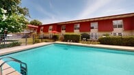 Located at 1334 Pascagoula St in Pascagoula, Mississippi, this single-level property features a well-appointed outdoor pool area that serves as a central recreational amenity. The centerpiece is a generously sized, crystal-clear swimming pool with a classic rectangular design, complemented by a wide surrounding deck finished in warm-toned pavers. The deck provides ample space for sunbathing and relaxation, furnished with reclining lounge chairs and additional seating arrangements, including a table and chairs shaded by a red umbrella.

A charming white cabana structure with French doors and a pitched roof sits at one corner of the pool area, offering a private retreat or utility space. The entire pool area is enclosed by a sturdy black iron fence, ensuring both safety and a defined sense of space. Lush tropical and subtropical landscaping, including palm trees, ornamental shrubs, and mature shade trees, frames the perimeter, creating a verdant, resort-like atmosphere that is well-suited to the Gulf Coast climate.

The adjacent building, clad in bold red siding with cream-colored trim, provides an attractive architectural backdrop. Large windows punctuate the facade, adding visual interest. The overall ambiance is inviting and well-maintained, blending practical amenities with vibrant, colorful surroundings ideal for outdoor leisure.