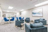 Located at 2 Quebec Street in Guelph, Ontario, this thoughtfully designed communal space serves as both a breakroom and lounge area, offering a comfortable and contemporary environment for relaxation and informal gatherings. The single-floor layout is efficiently organized into distinct functional zones that flow seamlessly into one another.

The kitchen area anchors one end of the space, equipped with a stainless steel refrigerator, microwave, and ample white cabinetry. A central white island with grey upholstered bar stools provides a casual spot for quick meals or conversation. Decorative wall accents and small green plants add a welcoming touch.

The dining zone features two round white tables surrounded by bold royal blue velvet chairs with chrome bases, creating a vibrant contrast against the neutral tones of the room. A textured white brick accent wall runs along one side, adding architectural character and visual warmth throughout the space.

The lounge area is furnished with plush grey velvet armchairs accented with gold legs, arranged around a metallic hourglass side table. A wall-mounted flat-screen TV and a landscape painting complete the relaxed ambiance. Patterned low-pile carpeting, a drop ceiling with recessed light panels, and taupe double doors with glass inserts round out this polished, functional, and inviting space.