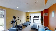 This fitness center offers a straightforward, functional space designed for cardiovascular exercise. The room features warm yellow walls accented by a bold red wall near the window, creating an energetic atmosphere suited to physical activity. A textured white ceiling with recessed lighting ensures the space is well-lit, complemented by natural light streaming in through a large window dressed with vertical blinds. The window also provides a view of greenery outside, adding a refreshing element to workouts. The carpeted flooring provides a cushioned surface underfoot. The space is equipped with two treadmills and an elliptical cross-trainer, offering residents a focused selection of cardio equipment. A potted plant near the window adds a touch of natural décor, softening the utilitarian feel of the room. The overall layout is efficient, positioning the equipment to maximize use of the available floor space while maintaining ease of movement between machines.