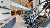 This professionally designed fitness center delivers a comprehensive workout environment across a spacious, open floor plan. Light hardwood flooring runs throughout, creating a clean, modern aesthetic that contrasts beautifully with exposed brick accent walls and warm, layered lighting from pendant fixtures and recessed ceiling lights. A dramatic wall of floor-to-ceiling windows floods the cardio zone with abundant natural light and outdoor views, creating an energizing atmosphere for treadmill and elliptical workouts. The main floor houses an extensive array of equipment, including treadmills, ellipticals, stair climbers, rowing machines, and a spin bike with an interactive display. A dedicated strength training zone features a multi-function rack, weight bench, and additional resistance equipment, anchored by a striking nature-themed mural that adds artistic depth to the space. A stylish staircase with cable railings leads to an upper mezzanine level, expanding the usable workout area and providing elevated views of the main floor. Additional amenities include a restroom, a wet bar or refreshment station with dark cabinetry, and ample open floor space for stretching or functional training. The overall ambiance is modern yet warm, blending industrial and boutique-gym sensibilities into a cohesive, high-end fitness environment.