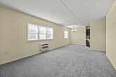 This single-floor unit offers a thoughtfully arranged layout that balances open living spaces with functional design. The main living area is generously proportioned, finished with neutral-toned carpet and warm cream-colored walls that create a bright, welcoming atmosphere. Multiple windows fitted with horizontal blinds allow natural light to fill the room, while a wall-mounted HVAC unit provides climate control. An elegant chandelier adds a refined touch to the dining or living zone.

The kitchen is a standout feature, showcasing rich dark-stained cabinetry with shaker-style doors, contrasting white quartz countertops, and a classic white subway tile backsplash. Stainless steel appliances — including a refrigerator, range, and built-in microwave — complete the modern aesthetic. Wood-look flooring grounds the space, and a peninsula counter with bracket-supported overhang provides a convenient casual dining or prep area, seamlessly connecting the kitchen to the main living space.

A hallway extends from the kitchen toward the bedroom, which is carpeted and finished in the same soft cream palette. The bedroom includes a paneled closet with ample storage. The overall layout, as seen in the dollhouse view, is compact yet efficient, making excellent use of every area within the unit.