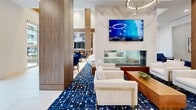 This sophisticated single-floor amenity space is designed with a contemporary aesthetic, blending clean architectural lines with warm, curated finishes. The main lounge area commands attention with its elevated ceiling, a striking dual-ring chandelier with cascading crystal accents, and large-format porcelain tile flooring that extends throughout. A bold navy and white geometric area rug anchors a plush seating arrangement centered around a sleek see-through fireplace, topped by a wall-mounted flat-screen TV set against a dramatic marble-look stone surround. Rich walnut wood paneling and accent columns add warmth to the bright, white palette.

A dedicated dining area features a long white communal table paired with cognac leather chairs, while the adjoining kitchenette showcases a white quartz island with walnut base, gold pendant lighting, brass fixtures, and a chevron-patterned tile backsplash wall. Beverage and coffee service equipment is neatly integrated into the station.

Private meeting and flex rooms enclosed by glass partitions provide intimate spaces for focused work or small gatherings, furnished with blue mid-century modern seating and bistro-style tables. A quiet alcove nook with navy lounge chairs and curated artwork offers an additional retreat. Floor-to-ceiling windows allow ample natural light and views to the surrounding exterior, enhancing the open, welcoming ambiance of the entire space.