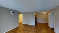 This single-story space presents a clean, functional layout with an open-concept design that connects the main living area to the kitchen. The flooring throughout is warm-toned hardwood-style planks, adding a sense of continuity and warmth to the interior. Neutral gray walls provide a versatile backdrop well-suited to a variety of décor styles.

The living area is spacious and unobstructed, benefiting from a wall-mounted HVAC unit that provides climate control. A textured popcorn ceiling runs throughout, adding subtle dimension overhead. White baseboards offer a crisp contrast against the gray walls and wood flooring.

Toward the rear of the space, a partial dividing wall separates the living area from the kitchen zone, where cabinetry and a stainless steel refrigerator are visible. A ceiling light fixture illuminates this area. An adjacent doorway leads to what appears to be a utility or laundry room, featuring bright overhead lighting and shelving. The overall ambiance is neutral and move-in ready, offering a practical and straightforward living environment.