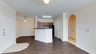 This single-floor space showcases an open-concept layout that seamlessly connects the living area to the kitchen, creating a spacious and functional flow. The kitchen is equipped with medium-toned wood cabinetry, black appliances including a refrigerator and range with overhead ventilation, and a dark-countertop peninsula that serves as both a workspace and a casual dining or gathering spot. Crown molding runs along the ceiling, adding a refined architectural detail throughout the main living area. The flooring consists of rich, dark hardwood-style planks that extend across the open space, providing a warm and grounded aesthetic. Neutral beige walls keep the interior feeling bright and versatile. An elegant arched doorway on one side of the space adds a classic architectural touch, leading to additional areas of the unit. A pendant light fixture in the dining area and overhead fluorescent lighting in the kitchen ensure ample illumination. The design balances everyday practicality with subtle traditional details, making the space both comfortable and visually cohesive.