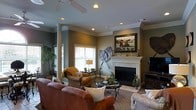This single-floor living space is thoughtfully designed with a blend of traditional elegance and comfortable functionality. The main living area serves as the focal point of the home, centered around a classic white-mantled fireplace adorned with decorative accents, including candle holders and a framed landscape painting above. Decorative wall art in the form of oversized leaf sculptures flanks the mantle, adding an artistic touch to the space.

The room features a decorative column detail that subtly defines the living area while maintaining an open feel. Crown molding runs along the perimeter of the ceiling, adding architectural refinement. Recessed lighting and a ceiling fan work together to provide both ambiance and practical illumination throughout the space.

Furnishings include rich leather sofas alongside upholstered accent chairs in warm, earthy tones, creating a welcoming and layered seating arrangement. A dark glass-top coffee table anchors the seating area, complemented by a neutral area rug. Natural light filters in through a window with white blinds, while potted indoor plants add life and greenery. An entertainment unit with a flat-screen television completes the room's functional layout.