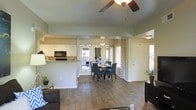 This single-floor residence showcases an open-concept layout that connects the living room, dining area, and kitchen in one cohesive, flowing space. Warm wood-style flooring extends throughout, lending a sense of continuity and warmth across the main living areas.

The living room features vaulted ceilings accentuated by a ceiling fan, adding both visual height and practical airflow to the space. Natural light filters in through multiple windows dressed with patterned curtains, brightening the neutral sage-toned walls.

The dining area comfortably accommodates a round table and chairs, positioned near a window bay that enhances the sense of openness. A pendant light fixture above the dining table adds a focused, refined touch.

The kitchen is defined by white cabinetry, a breakfast bar peninsula with granite-style countertops, and black appliances including a range and microwave. The peninsula serves as a natural divider between the kitchen and the living areas while maintaining the open feel.

Overall, the space offers a clean, functional design with cohesive finishes, making it both inviting and practical for everyday living.