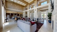 Located on Harvey Road in College Station, Texas, this single-story clubhouse facility is designed to serve as a premier communal gathering space. The architecture is immediately distinguished by its dramatic double-height ceilings, supported by structural columns and accented with coffered detailing and clerestory windows that flood the interior with abundant natural light throughout the day.

The main lounge area is generously proportioned, featuring light travertine-style tile flooring, elegant chandelier lighting, and comfortable seating arrangements that include plush sofas accented with bold crimson throw pillows. Floor-to-ceiling glass doors and expansive windows along the rear wall frame direct views of an outdoor pool area, seamlessly connecting the interior to the exterior amenity space. Sheer white drapery softens the abundant natural light while maintaining the open, airy atmosphere.

An adjacent game and social area provides a full-sized billiards table surrounded by casual seating clusters, making it an ideal space for recreation and socializing. The kitchen is well-appointed with rich dark wood cabinetry, stone tile backsplash, dark countertops, and stainless appliances, designed to support events and gatherings. The entry foyer features wainscoting detail and dual sets of glass-paned doors, providing a welcoming and bright arrival experience throughout this thoughtfully designed community facility.