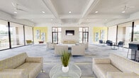 Located at 9310 Cherry Hill Road in College Park, Maryland, this single-floor communal space offers a generously sized, open-concept layout designed for both relaxation and collaboration. The interior is defined by its clean, contemporary aesthetic, featuring crisp white walls, coffered ceilings with recessed lighting, and ceiling fans that enhance air circulation throughout the space.

The patterned carpet—a stylish blend of gray, white, and yellow accents—runs throughout, tying the design together cohesively. A large lounge area is furnished with neutral-toned sofas and armchairs arranged around a central coffee table, creating an inviting atmosphere for social gatherings or quiet downtime. Adjacent to the lounge, a dedicated dining and meeting area offers table seating with modern chairs, ideal for group work or casual meals.

Floor-to-ceiling glass walls and sliding doors open directly onto an outdoor balcony, flooding the interior with abundant natural light and providing sweeping views of the surrounding greenery. Black-framed glass partitions and doors add a sophisticated architectural contrast to the bright white interiors. A centrally positioned service counter serves as a functional anchor point within the space. Colorful abstract artwork adorns the yellow accent wall, adding a vibrant, curated touch to the overall ambiance.