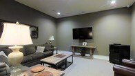 This single-floor space centers around a well-appointed living area designed with a cohesive, contemporary aesthetic. The room is finished with deep olive-gray walls that lend a warm, sophisticated ambiance, complemented by light neutral carpeting that softens the overall palette. Recessed ceiling lighting provides focused illumination throughout the space, creating a comfortable and functional environment.

A wall-mounted flat-screen television serves as the room's focal point, positioned above a rustic wooden console table with ornate baluster-style legs, adding a classic touch to the otherwise modern setting. Seating is anchored by a dark upholstered sofa adorned with accent pillows, paired with a reclaimed wood and metal coffee table that introduces an industrial-rustic element. A decorative round mirror with an ornate frame adds visual interest to one wall, while a classic table lamp provides warm ambient lighting.

The layout is open and well-proportioned, offering a comfortable flow through the space. Thoughtful décor details, including potted greenery and woven decorative accents, contribute to a grounded and inviting atmosphere throughout.