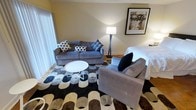 This single-floor space combines living and sleeping areas into one cohesive, efficiently designed layout. The living area serves as the centerpiece, featuring a grey upholstered sofa adorned with patterned throw pillows in black and white, complemented by a matching grey armchair with a navy accent pillow. A round white coffee table anchors the seating arrangement atop a bold geometric area rug in tones of black, white, and brown, adding visual interest to the flooring. A tall floor lamp provides warm ambient lighting, enhancing the room's inviting atmosphere. Neutral-toned walls create a calm backdrop, while a framed botanical art piece adds a decorative focal point. The sleeping area transitions naturally from the living space, with a neatly made bed visible in the background. Sheer vertical blinds along one wall allow for adjustable natural light. The overall ambiance is modern yet comfortable, with a cohesive color palette of grey, navy, and neutral tones throughout, creating a practical and stylish environment suited for both relaxation and rest.