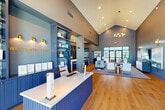 Located at 4400 Chemin Metairie Pkwy in Youngsville, Louisiana, the Sweetwater Point leasing office is a thoughtfully designed single-floor space that blends professional functionality with upscale interior aesthetics. The entrance greets visitors through floor-to-ceiling glass doors framed in sleek black steel, flooding the foyer with natural light and offering open views of the surrounding landscape. Soaring vaulted ceilings define the main reception area, where a bold navy blue fluted reception desk serves as the focal centerpiece, complemented by a stunning cascading gold chandelier overhead. Built-in shelving with decorative tiling provides an elegant backdrop behind the front desk. The open living lounge area features warm hardwood flooring, curated artwork, contemporary seating arrangements, and large windows that maintain a bright, airy atmosphere. A fully appointed kitchen includes an expansive white quartz island with bar seating, dark navy cabinetry, patterned tile backsplash, stainless steel appliances, and brass pendant lighting. A cohesive blue-and-gold color palette flows throughout the space, creating a polished, welcoming ambiance that reflects the identity of the Sweetwater Point community.