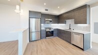This single-floor unit showcases a contemporary design with a cohesive aesthetic throughout. The kitchen is a standout feature, equipped with full stainless steel appliances including a gas range, built-in microwave, refrigerator, and dishwasher. Dark wood-textured cabinetry extends to the ceiling, contrasting elegantly with a white quartz countertop and a white subway tile backsplash with under-cabinet lighting. Light wood-style flooring runs throughout the main living areas, adding warmth to the modern palette.

The bedrooms are bright and airy, benefiting from large sliding windows that frame open city and neighborhood views. Each room is outfitted with a ceiling fan and connects to a walk-in closet with built-in shelving, offering generous storage. Crisp white walls and light flooring contribute to an open, clean atmosphere.

The bathroom is finished with large-format marble-look porcelain tile flooring and features a combined tub and shower enclosed by sliding glass doors. White horizontal tile lines the shower surround, while a floating vanity with dark wood cabinetry and an LED-backlit mirror completes the space. Recessed built-in niches provide additional storage within the shower area.