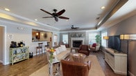 The Vinings Boulevard Clubhouse in O'Fallon, Missouri, is a thoughtfully designed single-floor community facility that combines comfort and functionality across several distinct spaces. The main lounge area is a welcoming gathering space, highlighted by a tray ceiling with crown molding, dual ceiling fans, and recessed lighting. A stone-surround fireplace anchors one wall, flanked by decorative star mirrors, while large windows with plantation shutters allow generous natural light throughout. Rich wood-look flooring adds warmth, and comfortable seating arrangements make the space ideal for casual socializing or relaxation. A flat-screen TV and entertainment console offer additional amenities for residents. Adjacent to the lounge is a well-appointed kitchen featuring warm wood cabinetry, granite countertops, stainless steel appliances, a subway tile backsplash, and a breakfast bar with pendant lighting — perfect for hosting community events. The conference room is a polished, professional setting centered around a large cherry wood table beneath a geometric chandelier, with a bay window providing a bright, airy atmosphere. A dedicated business center offers built-in workstations with granite surfaces, computer access, and printing capabilities. Together, these spaces create a versatile and inviting clubhouse suited for both social and professional needs.
