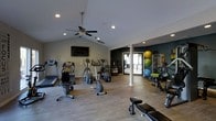 This fitness center offers a thoughtfully designed workout space spread across a single floor, combining functionality with a welcoming atmosphere. The room features vaulted ceilings accented by a ceiling fan and recessed lighting, creating an airy, open feel throughout. Light wood-style flooring runs the length of the space, contributing to a clean and modern aesthetic.

The cardio zone is prominently featured along one side of the room, housing multiple treadmills, elliptical trainers, and a stationary bike, providing a comprehensive selection of aerobic equipment. A wall-mounted flat-screen television is positioned nearby, ideal for entertainment during workouts.

A dedicated functional training area occupies the far corner, highlighted by a dark accent wall that adds a dynamic visual contrast. This zone includes a rack of stability balls in various sizes, resistance accessories, resistance bands, and a weight bench, catering to strength and flexibility training.

A structural column divides the space naturally, while an adjacent windowed area allows natural light to filter in, enhancing the overall brightness. The layout balances open floor space with organized equipment placement, supporting a variety of fitness routines in a comfortable environment.