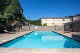 This outdoor community amenity space in Phenix City, Alabama offers a well-appointed gathering area designed for relaxation and recreation. At its centerpiece is a generously sized swimming pool with clear, blue water, surrounded by a broad concrete pool deck that provides ample room for lounge chairs and sunbathing. The pool area is enclosed by a wooden privacy fence, with mature trees lining the perimeter, offering natural shade and a lush backdrop under the open Alabama sky.

Adjacent to the pool is a covered pavilion supported by sturdy, rustic brick columns that lend a classic architectural character to the space. The pavilion features a wide, white-paneled roof that provides sheltered seating and includes both a picnic table and lounge chairs, making it an ideal spot for outdoor gatherings regardless of the weather. A concrete patio connects the pavilion seamlessly to the surrounding areas.

Access to the community is controlled through a decorative wrought-iron gate framed by matching brick columns, providing both security and a welcoming entrance aesthetic. The combination of brick detailing, iron fencing, and manicured outdoor furnishings gives the space a cohesive and appealing visual identity. Overall, this outdoor area presents a comfortable, community-focused environment well-suited for leisure and socializing.