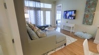 This single-floor living space offers a well-appointed interior centered around a comfortable, neutral-toned sectional sofa accented with decorative pillows. The living area features warm hardwood flooring throughout, grounding the space with a classic, inviting aesthetic. A patterned area rug defines the seating zone, complementing the overall neutral color palette. A wall-mounted flat-screen television is positioned opposite the sofa above a sleek white console table, creating a functional and visually balanced media area. Natural light fills the room through large windows dressed with patterned drapes, and a set of French doors provides access to an adjacent outdoor or interior space, enhancing the sense of openness. A small round coffee table with a geometric metal base adds a contemporary touch to the seating arrangement. A bench and floor lamp are also incorporated into the layout, contributing both style and practicality. The space presents a cohesive, modern design with clean lines and a light, airy ambiance suited for comfortable everyday living.