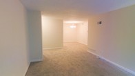 This single-floor space presents a clean, open layout connecting two main living areas. The living room is generously sized, finished with wall-to-wall beige carpeting that extends seamlessly into the adjoining dining area. Neutral beige walls and white baseboards throughout create a bright, cohesive aesthetic. A textured ceiling adds subtle visual interest overhead.

The transition into the dining area is defined by a partial wall, creating a sense of separation while maintaining an open, flowing feel between the two spaces. A chandelier serves as a focal point in the dining area, providing an elegant touch to the otherwise understated interior. A wall-mounted HVAC vent and visible baseboard heating registers indicate a functional climate control system integrated throughout.

Natural light filters softly into the space, complementing the warm tones of the walls and flooring. The layout offers versatile potential for furniture arrangement, making it well-suited for both relaxed living and dining use. Overall, the space conveys a simple, functional design with clean lines and a neutral palette that provides a welcoming, move-in ready environment.