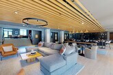 Located at 22 Mulberry Street in Worcester, MA, this impressive amenity floor spans a generous footprint and offers a thoughtfully designed collection of spaces for socializing, working, and recreation. The centerpiece is a expansive open-plan lounge and social hub, distinguished by striking slatted wood ceilings, a large circular pendant light, and plush sectional seating arranged around a wood coffee table. Adjacent to the lounge is a sleek kitchen and dining area featuring a dark-countered bar with stainless steel under-counter appliances, warm pendant lighting, and ample communal seating — all unified by the continuous wood slat ceiling detail. A separate living room showcases a sculptural wave ceiling, a vertical wood slat accent wall with a mounted flat-screen TV, and curated modern furnishings alongside a fireplace. Floor-to-ceiling windows flood the space with natural light and offer outdoor views. The fully equipped fitness center is a standout, boasting high ceilings, large industrial windows, cardio machines, strength equipment, and open training zones. A dedicated golf simulator room and private glass-walled offices round out the offering, making this a comprehensive, amenity-rich environment.