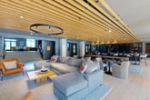 Located at 22 Mulberry Street in Worcester, MA, this impressive amenity floor spans a generous footprint and offers a thoughtfully designed collection of spaces for socializing, working, and recreation. The centerpiece is a expansive open-plan lounge and social hub, distinguished by striking slatted wood ceilings, a large circular pendant light, and plush sectional seating arranged around a wood coffee table. Adjacent to the lounge is a sleek kitchen and dining area featuring a dark-countered bar with stainless steel under-counter appliances, warm pendant lighting, and ample communal seating — all unified by the continuous wood slat ceiling detail. A separate living room showcases a sculptural wave ceiling, a vertical wood slat accent wall with a mounted flat-screen TV, and curated modern furnishings alongside a fireplace. Floor-to-ceiling windows flood the space with natural light and offer outdoor views. The fully equipped fitness center is a standout, boasting high ceilings, large industrial windows, cardio machines, strength equipment, and open training zones. A dedicated golf simulator room and private glass-walled offices round out the offering, making this a comprehensive, amenity-rich environment.