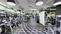 This contemporary fitness facility is thoughtfully designed to accommodate a wide range of workout styles and training needs, all within a single, expansive floor. The centerpiece of the space is a bold indoor sprint track, marked with numbered lanes and set against turf flooring, offering members a dedicated surface for sprinting, agility drills, and functional training.

Flanking the track on one side is a well-organized cardio zone, featuring a full row of treadmills and elliptical machines arranged for easy access and ample personal space. On the opposite side, a free weights area is equipped with a comprehensive selection of dumbbells and weight racks, catering to strength training enthusiasts of all levels.

Large mirrors line the walls throughout the facility, enhancing the sense of openness and allowing members to monitor their form during workouts. The interior is accented by vibrant green walls that add energy to the environment, complemented by bold, industrial-style pendant lights suspended from an exposed white ceiling with visible ductwork. The overall ambiance is clean, modern, and motivating, striking a balance between high-performance functionality and contemporary design aesthetics that make it an inviting space for fitness enthusiasts.