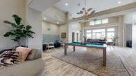 This single-story community clubhouse showcases a thoughtfully designed open layout with soaring ceilings and light-washed hardwood-style flooring throughout. The main gathering area serves as the social centerpiece, featuring a full-size pool table positioned atop a geometric area rug beneath a striking mid-century modern chandelier with brass accents and sculptural shades. Clerestory windows line the upper walls, flooding the space with abundant natural light and enhancing the sense of openness and volume.

Adjacent to the main area, a cozy lounge nook offers a teal upholstered booth-style banquette with patterned wall paneling, ideal for intimate conversation. Nearby, a separate seating zone features a large sectional sofa in teal fabric paired with a wood coffee table, anchored by a vibrant patterned area rug and oversized windows with exterior views.

A distinct fireplace lounge provides a more refined retreat, with a tiled surround fireplace, bold abstract artwork above, eclectic accent furniture, and a metallic gold oval coffee table set on a rich red Persian-style rug. A dedicated co-working area with computer stations rounds out the space, making this clubhouse equally suited for productivity, relaxation, and social engagement.