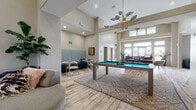 This single-story community clubhouse showcases a thoughtfully designed open layout with soaring ceilings and light-washed hardwood-style flooring throughout. The main gathering area serves as the social centerpiece, featuring a full-size pool table positioned atop a geometric area rug beneath a striking mid-century modern chandelier with brass accents and sculptural shades. Clerestory windows line the upper walls, flooding the space with abundant natural light and enhancing the sense of openness and volume.

Adjacent to the main area, a cozy lounge nook offers a teal upholstered booth-style banquette with patterned wall paneling, ideal for intimate conversation. Nearby, a separate seating zone features a large sectional sofa in teal fabric paired with a wood coffee table, anchored by a vibrant patterned area rug and oversized windows with exterior views.

A distinct fireplace lounge provides a more refined retreat, with a tiled surround fireplace, bold abstract artwork above, eclectic accent furniture, and a metallic gold oval coffee table set on a rich red Persian-style rug. A dedicated co-working area with computer stations rounds out the space, making this clubhouse equally suited for productivity, relaxation, and social engagement.