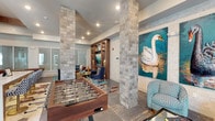 This single-floor amenity space is a thoughtfully designed community clubhouse that blends social functionality with bold, eclectic interior design. Distinctive natural stone columns anchor the open layout, creating defined zones while maintaining a sense of spaciousness throughout. The main lounge area features vibrant teal and gold accents, a geometric-patterned area rug, and a mix of plush seating including a tufted leather sofa and navy ottomans arranged around a built-in media wall with illuminated wood shelving and a flat-screen television. A wet bar area serves as a central gathering hub, complete with navy cabinetry, pendant globe lighting, a marble-topped counter with bar seating, and an under-counter refrigerator. The game room zone is equipped with a full-size foosball table and oversized Connect Four, offering ample entertainment options. A quieter lounge nook, adorned with large-scale swan paintings on a whitewashed brick wall, provides a relaxed retreat with a glass coffee table and patterned accent chairs. Floor-to-ceiling sliding glass doors flood the space with natural light and open to an outdoor courtyard, framed by dramatic teal and charcoal drapery panels. The overall ambiance is lively and curated, with art-forward décor and rich material contrasts throughout.