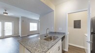 This single-floor residence showcases a thoughtfully designed open-concept layout that seamlessly connects the kitchen and living areas. The kitchen is outfitted with striking granite countertops featuring a blue-and-white pattern, bright white cabinetry with modern hardware, and a stainless steel sink paired with a brushed gold faucet. A dishwasher is integrated into the cabinetry for added convenience. The kitchen peninsula extends into the living space, creating a natural divide while maintaining an airy, connected feel. The living area benefits from natural light streaming through a window, and is equipped with a ceiling fan. Durable wood-look flooring runs throughout, complementing the neutral, light gray wall tones that lend a clean and contemporary aesthetic. The textured ceiling adds subtle visual interest while keeping the overall look cohesive. A doorway visible from the kitchen hints at additional rooms beyond, suggesting a functional and well-organized floor plan. The combination of modern finishes, practical fixtures, and an open layout makes this a comfortable and stylish living space suited for everyday living.