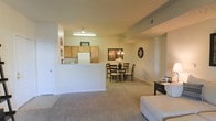 This single-floor space features an open-concept layout that seamlessly connects the living, dining, and kitchen areas into one cohesive environment. The living area is finished with neutral wall-to-wall carpeting, creating a warm and comfortable atmosphere. A half-wall partition defines the kitchen without fully closing it off, maintaining an open sightline throughout the space.

The kitchen features light wood cabinetry, white appliances including a full-size refrigerator, and overhead fluorescent lighting. The half-wall serves as a functional pass-through, making the kitchen feel integrated with the rest of the living space.

Adjacent to the kitchen, the dining area accommodates a four-person dark wood table set, lit by a decorative chandelier that adds a touch of elegance to the space. Neutral-toned walls throughout contribute to a clean, airy feel.

The living area, positioned at the front of the open floor plan, provides ample room for seating and everyday use. Recessed structural beams add subtle architectural detail to the ceiling, while HVAC vents indicate a central climate control system. The overall ambiance is casual, functional, and well-suited for comfortable daily living.