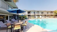 This two-story residential complex is thoughtfully designed around a central courtyard that serves as the heart of the property. The standout feature is a generously sized swimming pool with crystal-clear turquoise water, positioned at the center of the outdoor space and surrounded by a wide, sun-drenched pool deck. The deck is well-appointed with cushioned lounge chairs, teak-style dining tables, and vibrant blue market umbrellas, creating a resort-like atmosphere ideal for relaxation and outdoor entertaining.

The building's exterior presents a clean, mid-century modern aesthetic, with a smooth stucco facade in neutral tones accented by sage green trim and decorative iron railings. Upper-floor units feature private balconies that overlook the pool courtyard, offering residents direct views of the lush surroundings. Ground-floor units open directly onto the pool deck through sliding glass doors, seamlessly connecting interior and exterior living spaces.

Mature tropical landscaping enhances the ambiance throughout the property, with towering palm trees, large-leafed shrubs, potted plants, and dense greenery lining the perimeter walkways. Covered corridors run along the building's length, providing shaded passage between units. The overall atmosphere is bright, airy, and sun-soaked, characteristic of a warm Southern California setting.