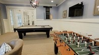 This single-floor game room is thoughtfully designed for recreation and social gatherings. The space features a classic black pool table and a full-sized foosball table as its centerpieces, complemented by a wall-mounted flat-screen television for additional entertainment. The room's aesthetic is defined by its crisp white wainscoting along the lower walls, contrasted by soft blue-gray upper walls that create a relaxed, inviting atmosphere. A textured neutral-toned carpet grounds the space and adds comfort underfoot. Overhead, a stylish spherical chandelier serves as a focal point, providing warm ambient lighting alongside recessed ceiling fixtures. Crown molding along the upper perimeter adds a refined architectural touch. French doors with glass panels lead to an adjacent room, allowing natural light to filter in and creating a sense of openness. A framed artwork piece and a track-lit display area on the wall contribute to the room's personalized character. The combination of classic design elements and recreational amenities makes this game room a versatile, well-balanced space suited for both casual play and relaxed socializing.