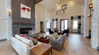 This single-floor space makes an immediate impression with its dramatically vaulted ceilings clad in white-painted wood paneling, lending an airy, lodge-inspired character throughout. Tall white brick columns frame the interior, reinforcing the vertical scale of the room and adding a refined architectural texture. A full-height stacked stone fireplace anchors one wall, serving as a striking focal point and contributing to the warm, inviting ambiance of the space.

Natural light flows generously through large windows and glass doors fitted with dark curtain panels, offering views of the surrounding outdoor environment. The flooring consists of wide-plank wood-look material that complements the neutral palette of the walls and furnishings. Seating is thoughtfully arranged throughout, featuring plush sofas, upholstered armchairs, and sleek accent chairs grouped around rustic wood coffee tables—creating multiple intimate conversation areas within the larger open plan.

Overhead, an industrial-style chandelier provides a modern contrast to the traditional stone and brick elements. Bold abstract artwork adorns the walls, adding color and visual interest. The overall atmosphere balances rustic warmth with contemporary design sensibility.