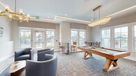 Located at 8263 NW Selvitz Rd in Port Saint Lucie, Florida, this single-floor community amenity space offers a well-appointed and versatile gathering environment. The layout flows seamlessly between a relaxed lounge area and an active recreation zone, making it ideal for both socializing and leisure.

The lounge area features a sleek linear fireplace set into a clean white wall, a wall-mounted TV flanked by coastal-themed artwork, and plush curved seating in a cool blue-gray tone. Warm gold-toned chandeliers add an elegant touch to the ambiance. Adjacent to the lounge, a wood-accented accent wall with integrated sconce lighting and a mounted TV creates a distinct focal point near the entryway corridor, which features light hardwood-style flooring.

The game area is anchored by a handsome wooden pool table with a statement pendant light overhead, surrounded by high-top bistro tables and bar-height seating. Multiple large windows along the perimeter flood the space with natural light and provide views of the outdoor pool and surrounding community. A cue rack along one wall and additional bistro seating complete the functional yet polished layout.

Throughout, textured carpet, tray ceilings with integrated LED cove lighting, and neutral warm tones create a cohesive, modern aesthetic that is both inviting and refined.