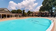 Located at 800 Walden Dr in Wilmington, North Carolina, this single-level outdoor community amenity space offers a well-appointed recreational environment designed for relaxation and social gatherings. The centerpiece is a generously sized swimming pool with a smooth curvilinear shape, surrounded by a wide sun deck lined with lounge chairs — ideal for enjoying the warm Carolina climate. A gated enclosure provides security and definition to the pool area, with a utility structure nearby serving the pool facility. Adjacent to the pool deck is a covered pergola structure supported by sturdy brick columns, featuring a built-in bar with high seating — a functional and attractive space for casual socializing. Additional open patio areas include picnic tables shaded by teal market umbrellas, providing comfortable spots for outdoor dining and gathering. A standout feature of the property is a white-painted octagonal gazebo fitted with a full wraparound sectional sofa and coffee table, creating an inviting open-air lounge. The gazebo's slatted design allows natural light and breezes to flow through while offering views of the surrounding mature greenery and lush landscaping. Together, these amenities form a cohesive and functional outdoor retreat well-suited for community use.