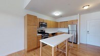 Located at Round Pointe Drive in Haverstraw, NY, this two-floor loft-style residence offers a thoughtfully designed layout that balances functionality with modern style. The main level features an open-concept kitchen equipped with a full suite of stainless steel appliances, including a gas range, over-the-range microwave, and refrigerator. Warm-toned wood cabinetry lines the walls, providing generous storage, while a white tile backsplash adds a clean, contemporary contrast. A central kitchen island with a white countertop and bar seating creates an ideal space for casual dining or entertaining. Hardwood flooring runs throughout, lending the space a bright and cohesive feel. The layout flows naturally between the kitchen and living areas, maximizing the open floor plan. With one and a half bathrooms across two levels, the residence offers practical convenience for daily living. The loft configuration adds architectural interest and a sense of verticality, making the space feel expansive. Clean white walls and recessed lighting contribute to the overall airy ambiance, creating a welcoming environment throughout the home.