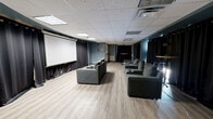 This single-floor screening and event space is designed to deliver an immersive entertainment experience within a well-appointed, intimate footprint. The room is defined by its large wall-mounted projection screen, which serves as the focal point and is flanked by floor-to-ceiling blackout curtains that line the perimeter walls. These heavy black drapes provide excellent light control and contribute to the room's theater-like atmosphere.

Seating consists of multiple sets of generously sized, upholstered sofas and lounge chairs with studded detailing and built-in cup holders, arranged in a viewing configuration facing the screen. The light wood laminate flooring provides a clean contrast to the dark curtains and teal-painted accent walls, lending the space a modern and polished aesthetic.

The drop tile ceiling houses recessed and flush-mounted lighting panels, along with a visible HVAC unit, ensuring comfortable climate control throughout. A tall bistro-style table with a pedestal base is positioned toward the rear of the room, offering a casual standing area suitable for networking or social gatherings. The overall design balances the functionality of a private screening room with the flexibility of a multipurpose event venue.