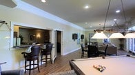 Located at 10210 Hillston Ridge Rd in Raleigh, this single-floor entertainment space is thoughtfully designed for hosting and leisure. The open layout connects several functional zones seamlessly, creating a cohesive environment ideal for gatherings. At the forefront is a full-sized pool table, positioned beneath warm ambient lighting that anchors the recreational area. Adjacent to it, a wet bar with dark leather upholstered bar stools and rich countertop finishes provides a sophisticated setting for entertaining guests. The bar area transitions into a separate, more intimate lounge space, partially enclosed to offer a distinct atmosphere. Continuing through the space, a dining area accommodates a sizeable table beneath a decorative pendant light fixture, complementing the overall aesthetic. Wall-mounted screens are strategically placed throughout, adding a modern, multimedia dimension to the space. Warm hardwood-style flooring runs consistently across the entire floor, tying the zones together. Crown molding along the ceiling adds a refined architectural touch, while recessed lighting ensures even illumination throughout. The neutral wall tones and classic furnishings contribute to an inviting, upscale ambiance.