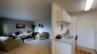 This single-story space showcases an open-concept design that seamlessly connects the kitchen and living area. The kitchen is equipped with white upper cabinetry offering ample storage, a countertop with an integrated sink, and a clean, efficient layout that keeps essentials within easy reach. A half-wall partition subtly defines the kitchen from the living space while maintaining an open, airy feel throughout.

The living area is generously sized, featuring neutral-toned carpet that adds warmth underfoot. Light gray walls provide a versatile backdrop, complemented by a textured ceiling that adds subtle visual interest. The room comfortably accommodates a full seating arrangement, including a sofa, armchair, and coffee table, demonstrating its functional scale.

Framed artwork adorns the walls, contributing to a welcoming ambiance. A floor lamp and decorative plants enhance the room's warmth. The overall design is practical and straightforward, with a neutral color palette throughout that creates a cohesive, calm atmosphere suitable for a variety of furnishing styles.