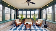 Located at 1508 Madison Ave in Memphis, Tennessee, this sunroom is a characterful and inviting retreat that masterfully blends rustic and classic design elements. The space is defined by its exposed brick walls, which lend warmth and historic character throughout. Bold, deep green window trim frames an expansive array of multi-pane windows on multiple walls, flooding the interior with abundant natural light and offering open views of the surrounding outdoor scenery.

The eye-catching blue-and-white checkered floor serves as a striking focal point, adding a playful yet timeless aesthetic to the room. Crown molding lines the ceiling, contributing a refined architectural detail, while a ceiling fan with a decorative light fixture provides both comfort and ambiance. String lights are strung along the upper trim, adding a warm, inviting glow.

The space is divided into two distinct seating zones: one featuring a rattan loveseat, matching armchairs, and a rattan coffee table arranged atop the patterned rug, and another more intimate area with a wrought iron bistro table and chairs set near an ornate glass-paneled entry door. Tall potted palms punctuate the space, enhancing its bright, airy atmosphere. A small wood-burning stove adds a cozy, functional touch, making this sunroom a versatile and welcoming space year-round.