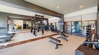 Located on Old Monrovia Road NW in Huntsville, Alabama, this fitness center offers a thoughtfully designed, single-floor layout that accommodates a wide range of workout needs. The space is organized into distinct functional zones, each serving a specific training purpose while maintaining an open, cohesive flow throughout.

The strength training area is anchored by a large multi-station cable machine and a full power rack system with barbells and weight plates, complemented by a selection of adjustable benches for free weight exercises. A dedicated dumbbell rack lines one wall, accompanied by floor-level storage for plyo boxes and accessories. Large mirrors span multiple walls, enhancing the sense of space and providing visual feedback during training.

The cardio zone features a row of treadmills and recumbent bikes, positioned along floor-to-ceiling windows that overlook an outdoor pool area and surrounding grounds, providing users with views and natural daylight. Roller shades offer adjustable light control throughout the day.

The interior design incorporates a neutral palette of white and gray walls accented by dark charcoal architectural banding, with light carpet flooring interrupted by warm wood-tone strips. Recessed lighting throughout ensures consistent illumination. Wall-mounted televisions are positioned across the space for convenience during workouts.