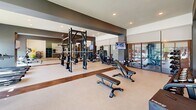 Located on Old Monrovia Road NW in Huntsville, Alabama, this fitness center offers a thoughtfully designed, single-floor layout that accommodates a wide range of workout needs. The space is organized into distinct functional zones, each serving a specific training purpose while maintaining an open, cohesive flow throughout.

The strength training area is anchored by a large multi-station cable machine and a full power rack system with barbells and weight plates, complemented by a selection of adjustable benches for free weight exercises. A dedicated dumbbell rack lines one wall, accompanied by floor-level storage for plyo boxes and accessories. Large mirrors span multiple walls, enhancing the sense of space and providing visual feedback during training.

The cardio zone features a row of treadmills and recumbent bikes, positioned along floor-to-ceiling windows that overlook an outdoor pool area and surrounding grounds, providing users with views and natural daylight. Roller shades offer adjustable light control throughout the day.

The interior design incorporates a neutral palette of white and gray walls accented by dark charcoal architectural banding, with light carpet flooring interrupted by warm wood-tone strips. Recessed lighting throughout ensures consistent illumination. Wall-mounted televisions are positioned across the space for convenience during workouts.