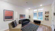 This single-floor space presents a clean, contemporary aesthetic centered around a well-appointed lounge area. The room features warm hardwood flooring that grounds the space, complemented by crisp white walls and a smooth white ceiling equipped with recessed lighting that provides even, ambient illumination throughout.

The seating area is anchored by a sleek black leather sofa with chrome accents, paired with a neutral-toned accent chair adorned with a patterned throw pillow. A bold black-and-white striped area rug defines the lounge zone, adding graphic visual interest to the hardwood floor beneath. A low black bench with metal legs serves as both a functional and decorative element near the window.

Wall art plays a prominent role in the room's character, with two framed pieces — one featuring bold red and black abstract strokes — adding color and personality to the otherwise neutral palette. A tall white cylindrical floor lamp provides additional warm lighting, while a potted tropical plant introduces a natural, organic element to the modern setting.

Large windows along one wall invite generous natural light into the space, brightening the interior and creating an open, airy atmosphere. The overall design is minimal yet refined, balancing modern furnishings with thoughtful decorative touches to create a comfortable and visually cohesive environment.