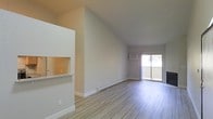 This single-floor space offers a clean, modern interior defined by soaring vaulted ceilings that lend an airy, expansive feel to the main living area. The open floor plan is anchored by light-toned vinyl plank flooring that runs throughout, complementing the neutral, warm-white wall palette for a cohesive and contemporary aesthetic.

A defining architectural feature is the pass-through window connecting the kitchen to the living area, framed with a granite-style countertop ledge — an ideal detail for casual dining or entertaining. The kitchen is partially visible through this opening, hinting at a well-integrated layout designed for ease of daily living.

The living area is generously proportioned, with natural light streaming in through a large sliding glass door that opens to a private balcony, offering a seamless indoor-outdoor connection. A wall-mounted air conditioning unit ensures year-round comfort. Baseboard heating runs along the lower walls, providing an additional climate control option.

The space is presented in a vacant, move-in-ready condition, allowing the architectural details — the dramatic ceiling pitch, clean lines, and thoughtful layout — to speak for themselves. Overall, the space exudes a bright, welcoming ambiance suited to a variety of living arrangements.